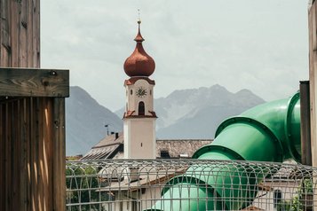 Eine grüne Röhrenrutsche auf dem Außenspielplatz im Familienhotel Tirolerhof mit Blick auf die Kirche und die Tiroler Bergkulisse