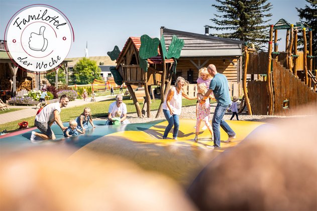 Familien auf einem großen Hüpfkissen auf einem Abenteuerspielplatz, Kinder und Erwachsene lachen und spielen vor Holz-Spielgeräten.