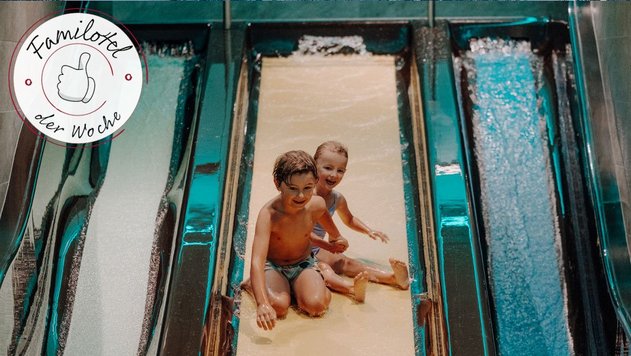 Zwei Kinder sitzen hintereinander auf einer breiten Wasserrutsche im Hallenbad und rutschen durch spritzendes Wasser nach unten; links oben steht „Familotel der Woche“.