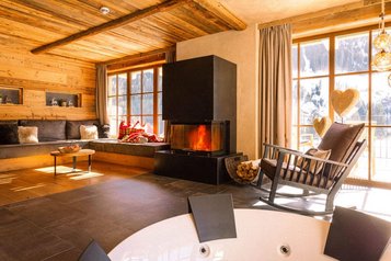 Helles Wohnzimmer im Alpura Retreat mit Kamin, großer Fensterfront, Holzwänden, gemütlicher Sitzecke und Blick auf verschneite Berge.