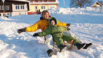 Mutter und Tochter sitzen auf einem Schlitten im Schnee.