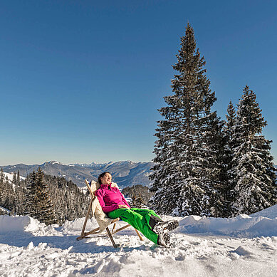 Eine Frau sitzt im Stuhl geniest die Sonne. Der Wald ist Schnee bedeckt.
