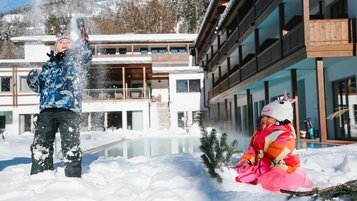 Zwei Kinder spielen im Schnee vor dem Familienhotel Das Bayrischzel mit beheiztem Außenpool, eines wirft Schnee in die Luft, das andere sitzt im Schnee neben einem kleinen Tannenzweig.