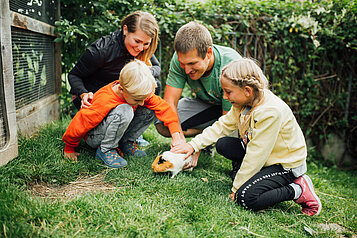 Familie mit zwei Kindern streichelt ein Meerschweinchen im Garten des Kinderhotels Sailer in Tirol.