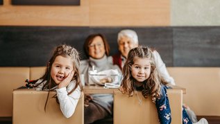 Zwei Mädchen lehnen lächelnd über eine Sitzbank, im Hintergrund sitzen die Großeltern am Restauranttisch im AIGO Welcome Family.