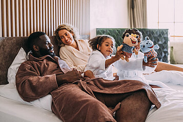 Eltern kuscheln mit ihren zwei Kindern im großen Bett im Familienhotel Post Family Resort im Salzburger Land.