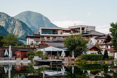 Sommeransicht des Familienhotels Post mit Blick auf den Naturbadeteich und die umliegenden Berge.
