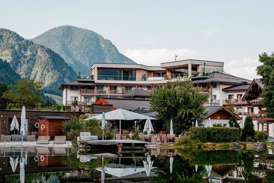Sommeransicht des Familienhotels Post mit Blick auf den Naturbadeteich und die umliegenden Berge.