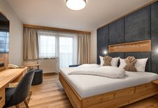 Modernes Doppelzimmer im Hotel Zauchenseehof mit großem Holzbett, dunkel gepolstertem Kopfteil, zwei dekorativ gefalteten Bademänteln auf den Kissen, hellem Holzfußboden, Schreibtisch und großen Fenstern mit Tageslicht.