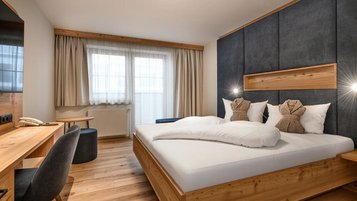 Modernes Doppelzimmer im Hotel Zauchenseehof mit großem Holzbett, dunkel gepolstertem Kopfteil, zwei dekorativ gefalteten Bademänteln auf den Kissen, hellem Holzfußboden, Schreibtisch und großen Fenstern mit Tageslicht.