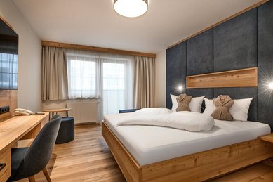 Modernes Doppelzimmer im Hotel Zauchenseehof mit großem Holzbett, dunkel gepolstertem Kopfteil, zwei dekorativ gefalteten Bademänteln auf den Kissen, hellem Holzfußboden, Schreibtisch und großen Fenstern mit Tageslicht.