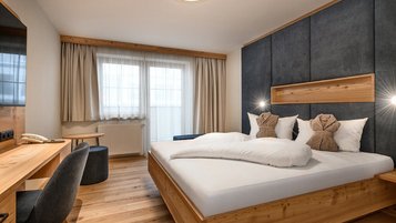 Modernes Doppelzimmer im Hotel Zauchenseehof mit großem Holzbett, dunkel gepolstertem Kopfteil, zwei dekorativ gefalteten Bademänteln auf den Kissen, hellem Holzfußboden, Schreibtisch und großen Fenstern mit Tageslicht.