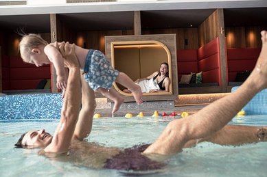 Ein Vater ist mit seinem Kind im Hotelpool und hebt in mit seinen Armen in die Luft, im Hintergrund liegt die Mutter gemütlich auf einer großen Liege.