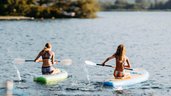 Zwei Kinder paddeln im Sommer auf dem See mit SUP-Boards beim Familienhotel Amiamo vor malerischer Bergkulisse.