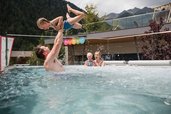 Vater hebt lachendes Kind in die Luft, während Mutter mit Baby im Arm im warmen Outdoor-Pool des Familienhotels Huber steht, im Hintergrund Berge und moderne Terrasse.