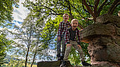 Wandern in der Rhön mit Kindern: Zwei Jungen klettern auf einem Felsen und lachen. Im Hintergrund sind Bäume zu sehen.