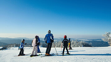 Eine Familie steht auf einer Skipiste mit ihren Skiern und genießt im Bayerischen Wald den Ausblick auf Mitterdorf im Winter.