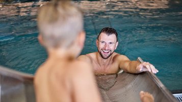 Junge rutscht die Wasserrutsche im Hallenbad im Familienhotel Amiamo in Zell am See hinunter und der Vater wartet lächelnd im Wasser auf ihn.