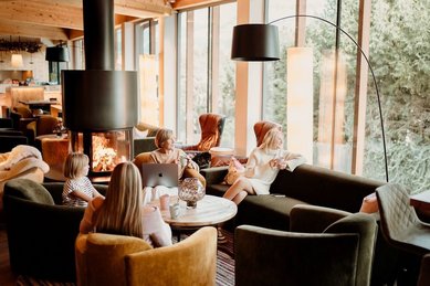 Gemütliche Lounge im Familienhotel Mia Alpina in Tirol mit Kamin, großen Fenstern, Sesseln und Gästen beim Entspannen.