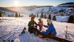Familie in Skiausrüstung sitzt im Schnee auf einer Bank, trinkt aus Thermobechern und blickt auf ein sonniges Wintertal mit verstreuten Häusern und verschneiten Hügeln.