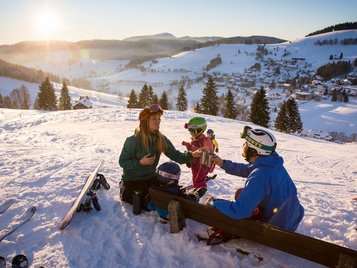 Familie in Skiausrüstung sitzt im Schnee auf einer Bank, trinkt aus Thermobechern und blickt auf ein sonniges Wintertal mit verstreuten Häusern und verschneiten Hügeln.