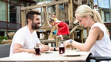 Familie beim Essen auf der Terrasse eines Hotels, währen die beiden Kinder auf einem Spielturm mit Eimer und Sand spielen.