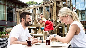 Familie beim Essen auf der Terrasse eines Hotels, währen die beiden Kinder auf einem Spielturm mit Eimer und Sand spielen.