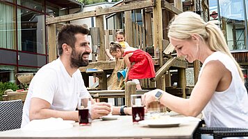 Familie beim Essen auf der Terrasse eines Hotels, währen die beiden Kinder auf einem Spielturm mit Eimer und Sand spielen.