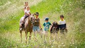 Kinder erleben gemeinsam mit einer Begleiterin einen Ponyritt über eine blühende Almwiese beim Familienhotel Bella Vista Südtirol.