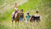 Kinder erleben gemeinsam mit einer Begleiterin einen Ponyritt über eine blühende Almwiese beim Familienhotel Bella Vista Südtirol.