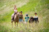 Kinder erleben gemeinsam mit einer Begleiterin einen Ponyritt über eine blühende Almwiese beim Familienhotel Bella Vista Südtirol.