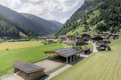 Mehrere Gebäude des Familienhotels Oberkarteis im Salzburger Land sind aus der Vogelperspektive mit einem teilweise überdachten Sandplatz zum Reiten vor Wiesen und Bergen zu sehen.