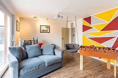Modern gestalteter Teenager-Raum im Familienhotel Deichkrone mit einem Tischfußball, bequemen Sofas und einer hängenden Ei-Stuhlschaukel, der zur Entspannung und Unterhaltung junger Gäste einlädt.