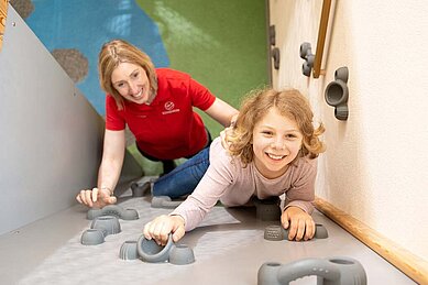 Mädchen klettert die Boulderwand im Happy-Club des Familienhotels Sonnenpark nach oben. Hinter ihr steht eine Kinderbetreuerin und passt auf sie auf.