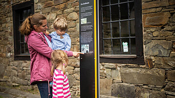 Mutter mit zwei Kindern steht vor einer Infosäule eines Museums.