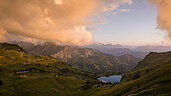 Ein wunderschönes Panorama mit Bergen, Landschaft und See im Allgäu. Die Wolken sind orange und die Berge grün.