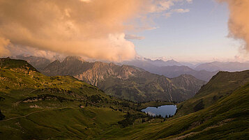 Ein wunderschönes Panorama mit Bergen, Landschaft und See im Allgäu. Die Wolken sind orange und die Berge grün.