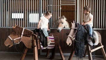 Zwei Kinder sitzen auf Holzpferdem und lachen. Hinter ihnen steht ein Pony in einer Box am Ponyhof. Der perfekte Ort für alle Pferdereunde: Urlaub im Elldus Resort im Erzgebirge