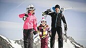 Familie mit Kind geht in Skiausrüstung und mit Skiern auf den Schultern in Richtung Skigebiet.