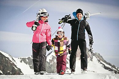 Familie mit Kind geht in Skiausrüstung und mit Skiern auf den Schultern in Richtung Skigebiet.