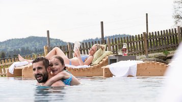 Familien Outdoorpool, Papa und tochter plantschen im Wasser. Mama liegt auf der Sonnenliege und liest ein Buch.
