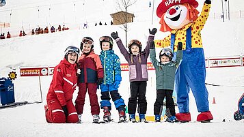 Einige Kinder bei einer Skischule jubeln mit dem Familotel-Maskottchen Happy