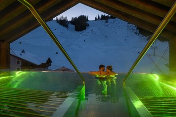 Paar genießt im beleuchteten Infinity-Pool des Zauchenseehof den Blick auf die abendliche, schneebedeckte Skipiste.