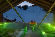 Paar genießt im beleuchteten Infinity-Pool des Zauchenseehof den Blick auf die abendliche, schneebedeckte Skipiste.