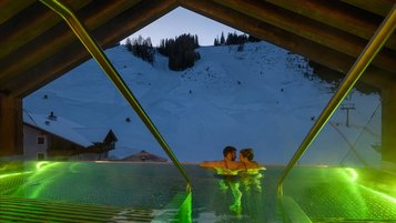Paar genießt im beleuchteten Infinity-Pool des Zauchenseehof den Blick auf die abendliche, schneebedeckte Skipiste.