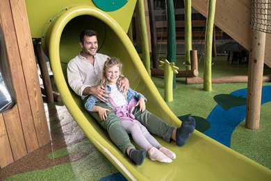 Vater und Tochter genießen eine fröhliche Rutschpartie im Indoor Spielbereich des Familienhotels mit natürlichem Design und viel Platz zum Toben