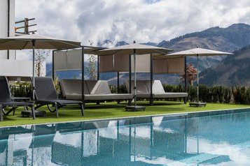 Poolbereich mit modernen Daybeds und Sonnenschirmen im Familienhotel Almfamilyhotel Scherer vor beeindruckender Bergkulisse.