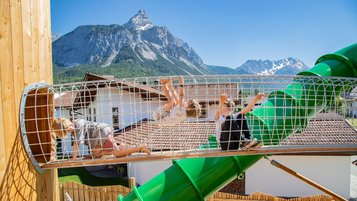 Kinder klettern auf einem Spielplatz mit grüner Röhrenrutsche vor alpiner Bergkulisse.
