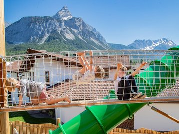 Kinder klettern auf einem Spielplatz mit grüner Röhrenrutsche vor alpiner Bergkulisse.