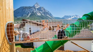 Kinder klettern auf einem Spielplatz mit grüner Röhrenrutsche vor alpiner Bergkulisse.